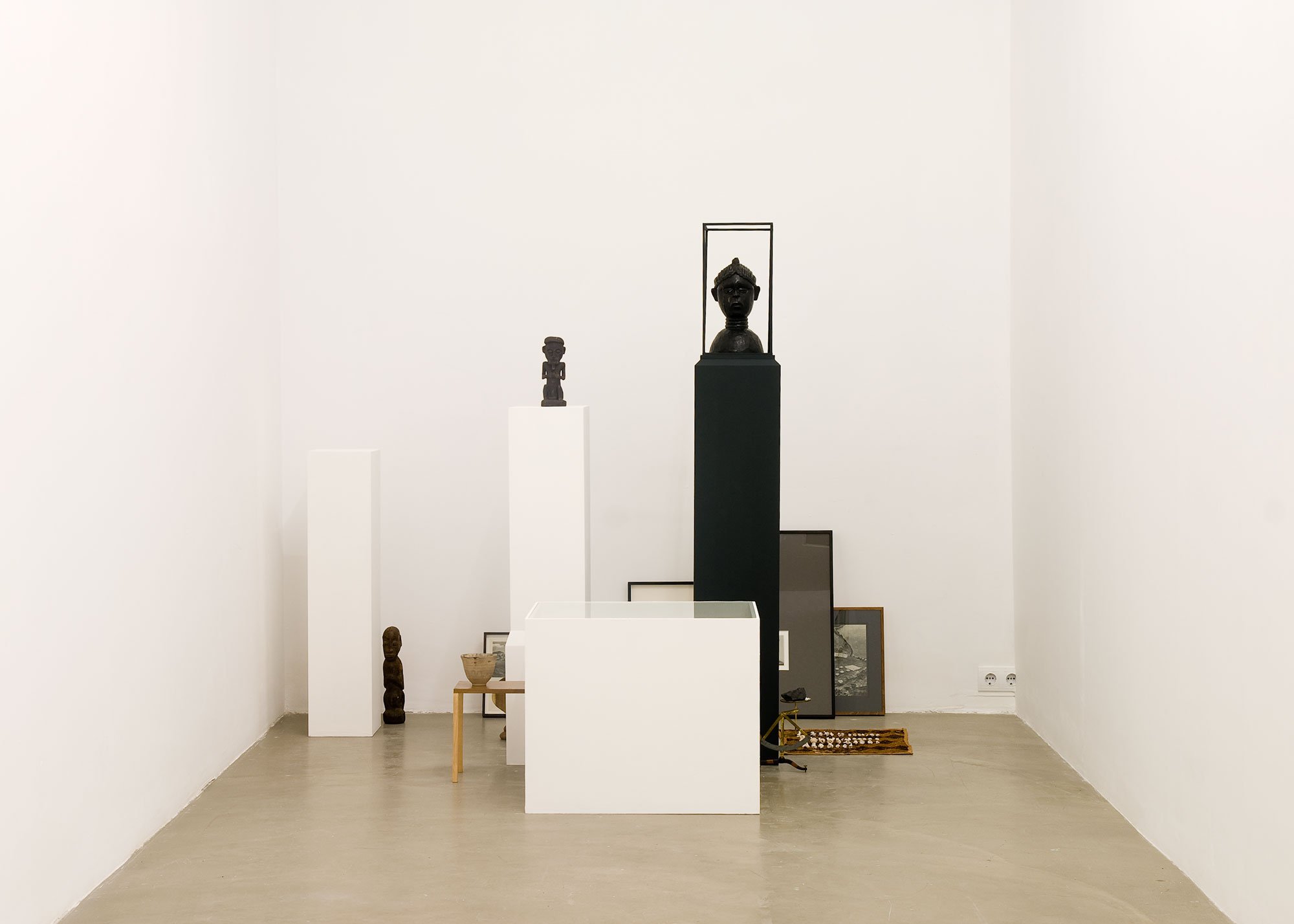 Haris Epaminonda, installation with three plinths, three African statuettes, a carpet with bones (49 pieces), a wooden statue of a cobra snake, an urn, a scale, nine frames on the floor and a vitrine that contains a Chinese book and an old stamp. Installation view, VOL. IV, Rodeo, Istanbul, 2009