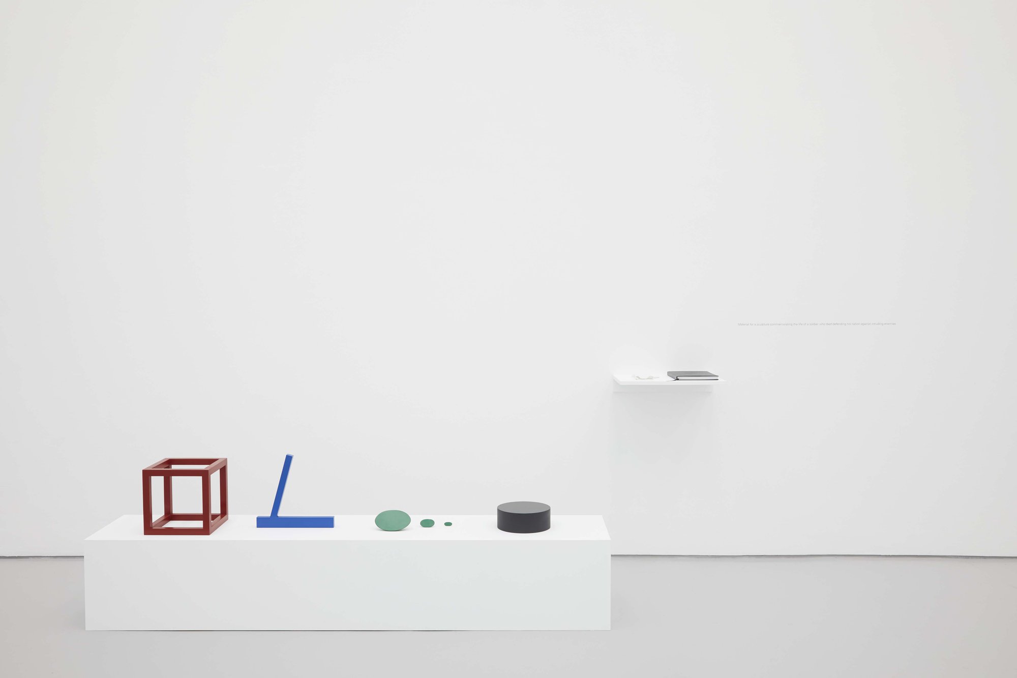 Iman Issa, Material for a sculpture commemorating the life of a soldier who died defending his nation against intruding enemies, four painted wooden sculptures, painted wooden plinth, painted wooden shelf, blank book with four inserts, vinyl text on wall, 2012