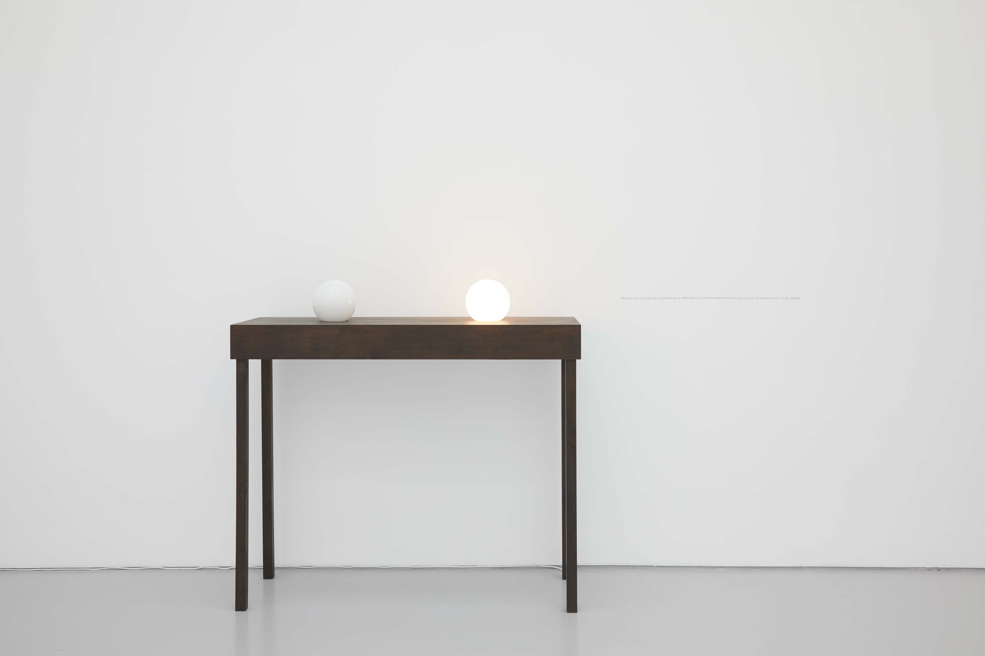 Iman Issa, Material for a sculpture proposed as an alternative to a monument that has become an embarrassment to its people, two light blubs, dark walnut plywood table, vinyl text on wall, 150 x 120 x 50 cm (59 x 47 1/4 x 19 2/3 in), 2010