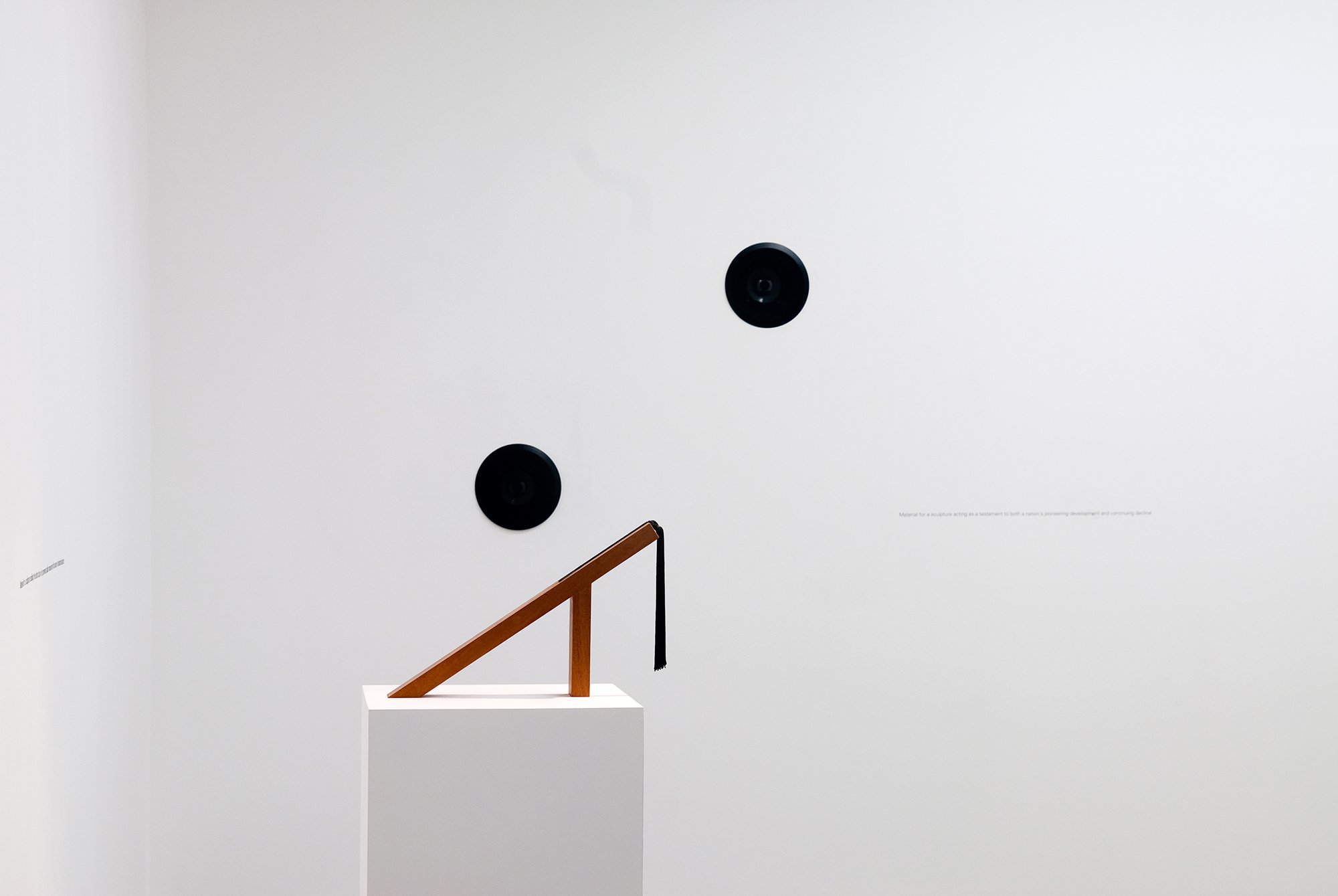 (On plinth) Iman Issa, Material for a sculpture recalling the destruction of a prominent public monument in the name of national resistance, mahogany sculpture with black tassel, white wooden pedestal, 142 x 49 cm, 64 points vinyl writing, 2010. (On wall) Iman Issa,  Material for a sculpture acting as a testament to both a nation’s pioneering development and continuing decline, sound installation, 10 sec. sound in 5 min., interval, 64 points vinyl writing, 2011. Installation view, Material, Rodeo, Istanbul, 2011