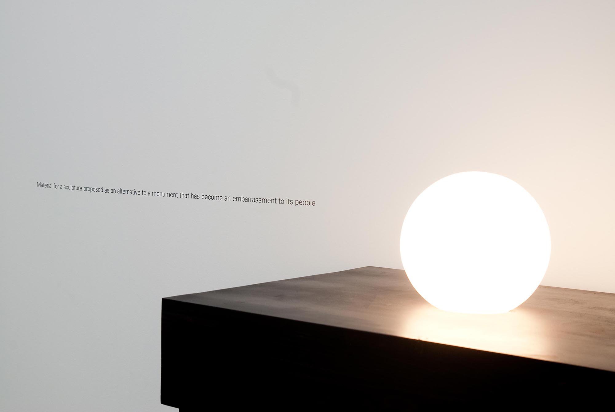 Iman Issa, Material for a sculpture presented as an alternative to a monument that has become an embarrassment to its people, detail, 2 light bulbs, dark walnut plywood table, 150 x 50 x 120 cm (59 x 19 3/4 x 47 1/4 in), light bulbs are synced in a loop, one lights up while the other slowly fades, 64 points vinyl writing, 2010